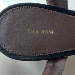 Peut inclure: Gros plan sur une semelle de sandale en cuir marron avec l'inscription "THE ROW" en lettres dor&eacute;es. La semelle int&eacute;rieure est entour&eacute;e d'une bordure noire et d'une bride en daim marron.