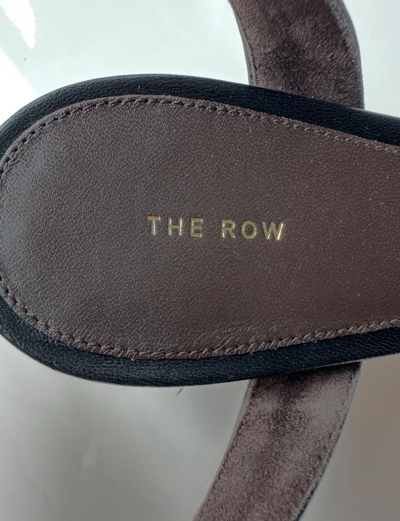 Peut inclure: Gros plan sur une semelle de sandale en cuir marron avec l'inscription "THE ROW" en lettres dor&eacute;es. La semelle int&eacute;rieure est entour&eacute;e d'une bordure noire et d'une bride en daim marron.