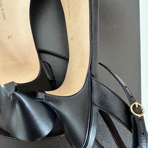 Peut inclure: Chaussures &agrave; talons en cuir noir avec brides de cheville et boucle dor&eacute;e. Les semelles beiges portent l'inscription "MADE IN ITALY" et la taille "41". Les chaussures sont sur une surface noire.
