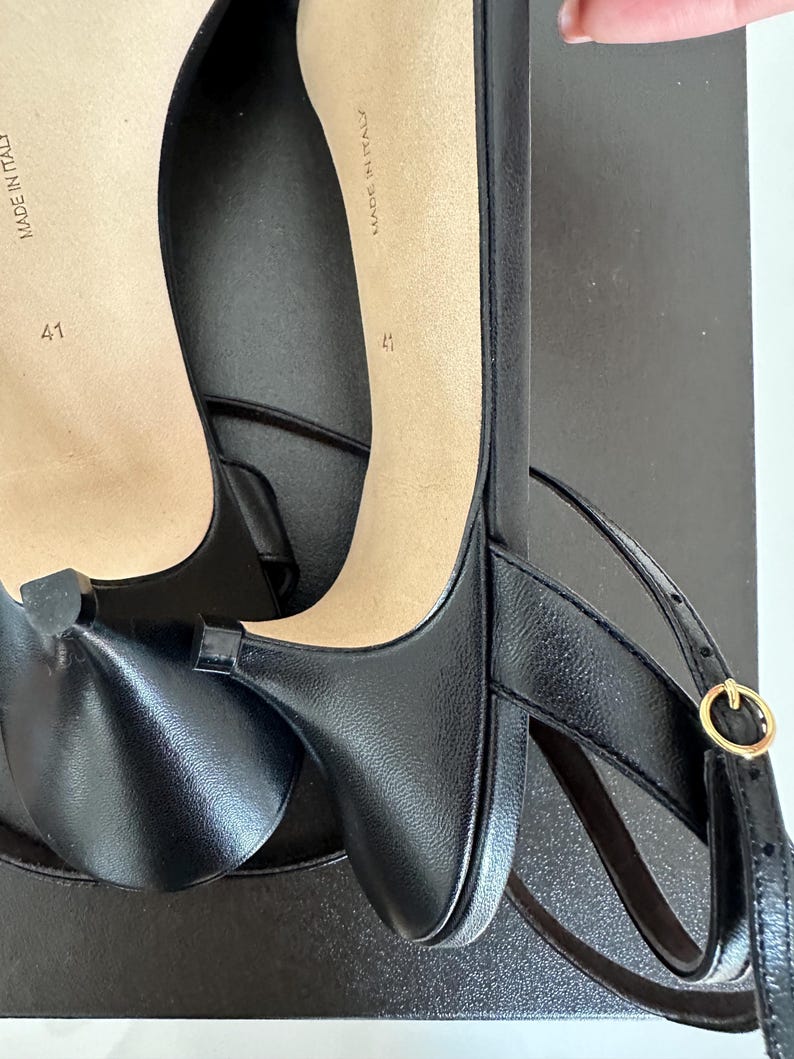 Peut inclure: Chaussures &agrave; talons en cuir noir avec brides de cheville et boucle dor&eacute;e. Les semelles beiges portent l'inscription "MADE IN ITALY" et la taille "41". Les chaussures sont sur une surface noire.