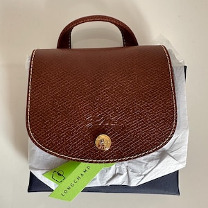 Puede incluir: Un bolso de mano de cuero marrón con asa superior y cierre de solapa. El bolso presenta costuras blancas y un cierre de botón dorado. Una etiqueta verde con la palabra "LONGCHAMP" está adjunta.