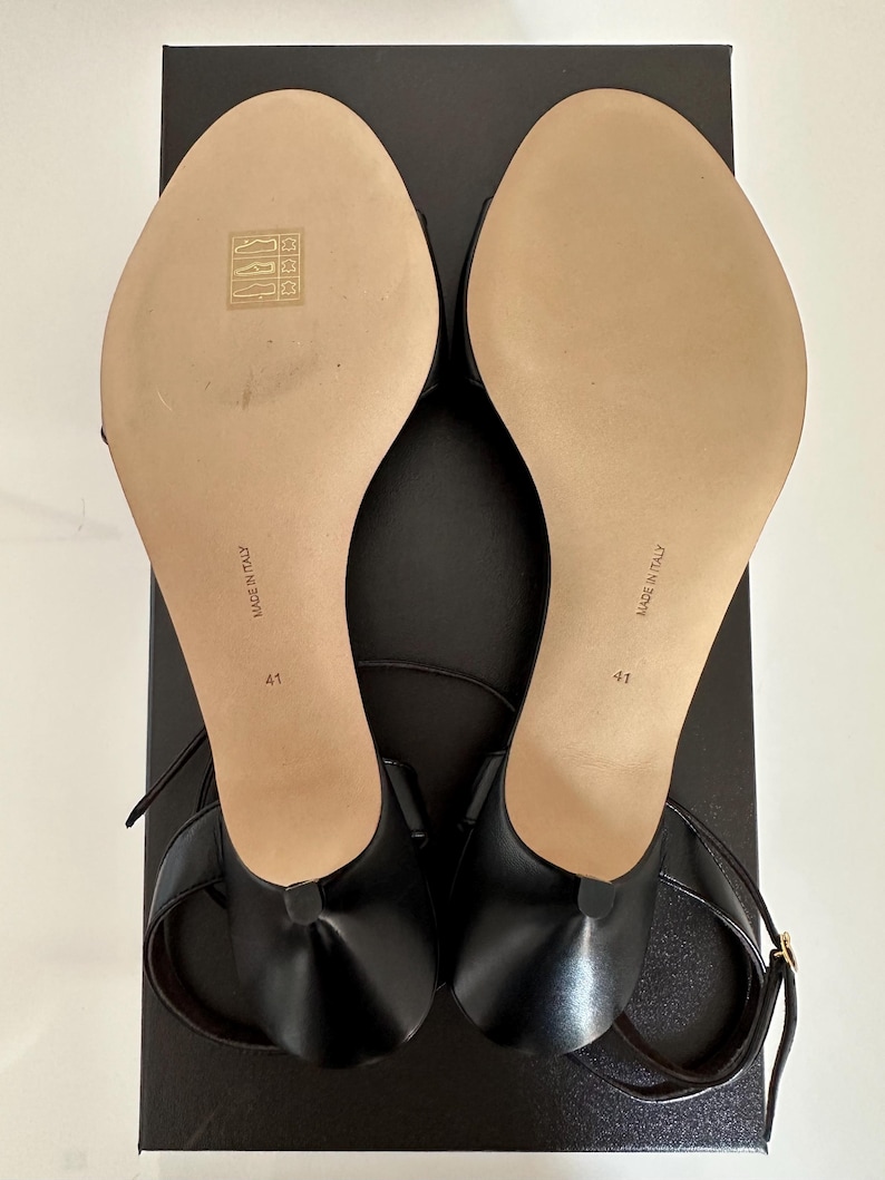 Peut inclure: Paire de sandales en cuir noir avec fines lani&egrave;res de cheville, taille 41, pos&eacute;es sur une bo&icirc;te noire. Les semelles beiges portent la mention "MADE IN ITALY". Les sandales ont un petit talon et un design minimaliste.