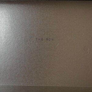 Peut inclure: Gros plan d'une surface marron textur&eacute;e avec le texte "THE ROW" au centre. La surface semble faire partie d'un conteneur ou d'un article similaire.
