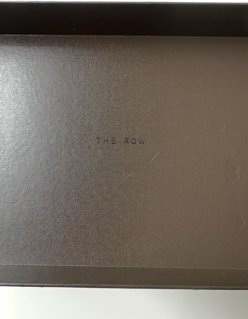 Peut inclure: Gros plan d'une surface marron textur&eacute;e avec le texte "THE ROW" au centre. La surface semble faire partie d'un conteneur ou d'un article similaire.