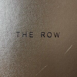 Peut inclure: Gros plan sur une surface marron textur&eacute;e avec l'inscription "THE ROW" en noir. La surface pr&eacute;sente un motif granuleux subtil et le texte est centr&eacute;.
