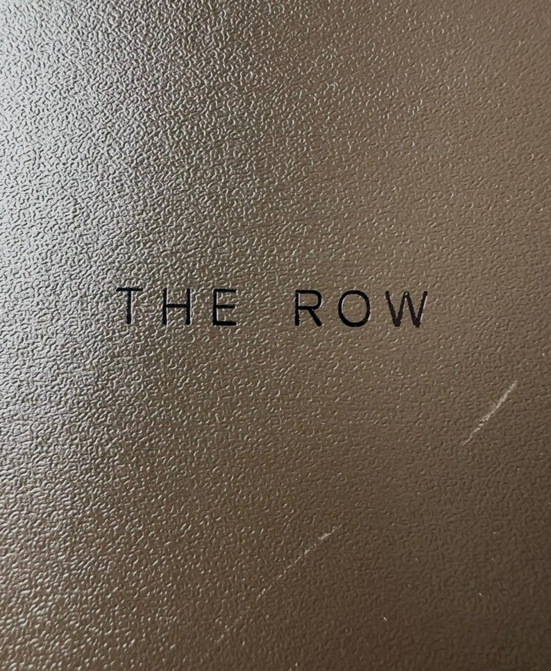 Peut inclure: Gros plan sur une surface marron textur&eacute;e avec l'inscription "THE ROW" en noir. La surface pr&eacute;sente un motif granuleux subtil et le texte est centr&eacute;.
