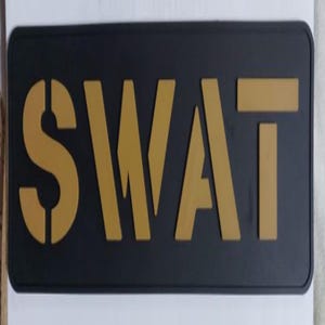 May include: A rectangular black sign with the word "SWAT" in gold stencil-style lettering. The letters are bold and stand out against the dark background. The sign has rounded corners.