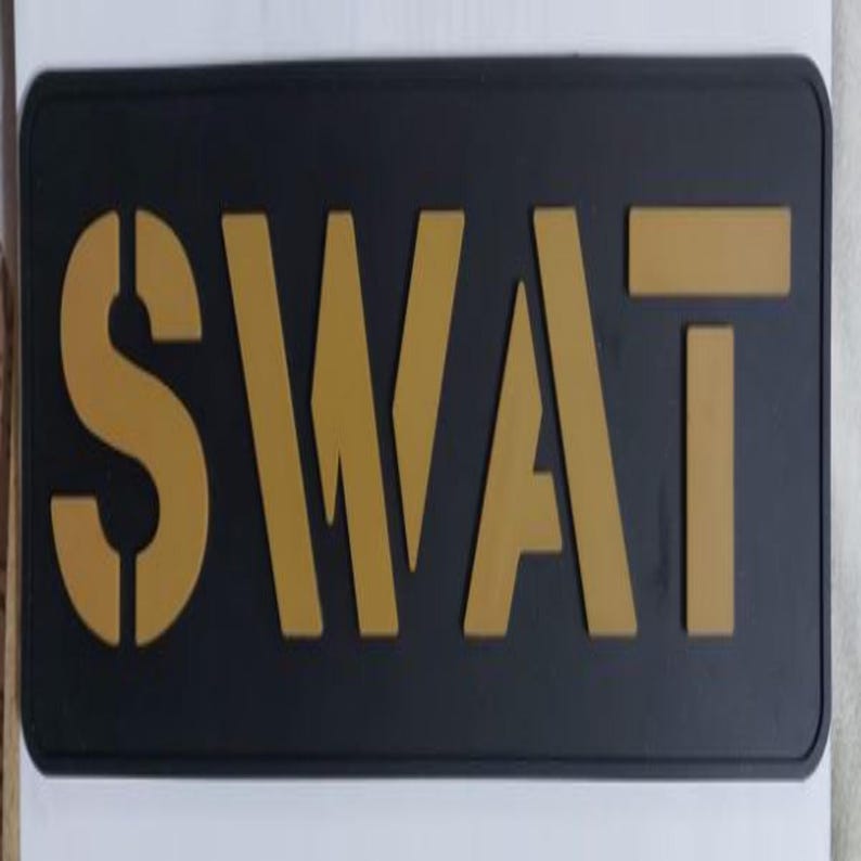 May include: A rectangular black sign with the word "SWAT" in gold stencil-style lettering. The letters are bold and stand out against the dark background. The sign has rounded corners.