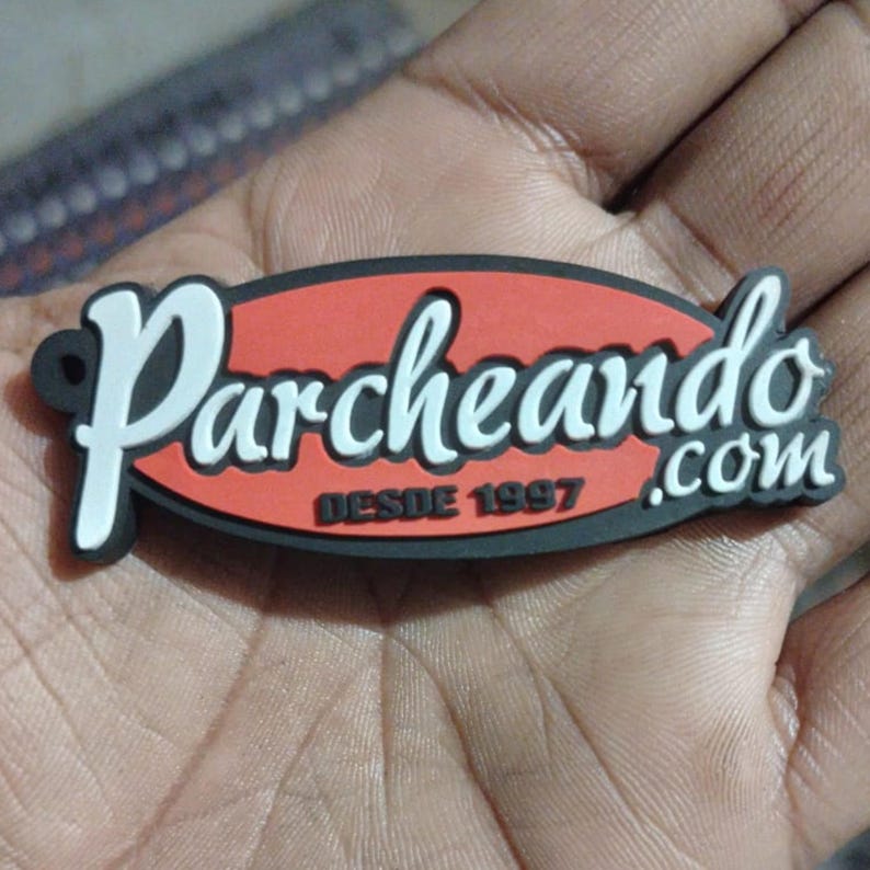 May include: A black, red, and white oval-shaped magnet with the word "Parcheando" in white script. The text "DESDE 1997" is below the word "Parcheando" in black text. The word ".com" is in white script. The magnet is held in a hand.