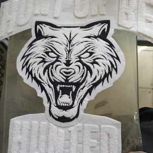 May include: A white and black embroidered wolf patch with the text "HUNT OR BE HUNTED". The wolf has an open mouth with visible teeth. The text is in a block letter style.