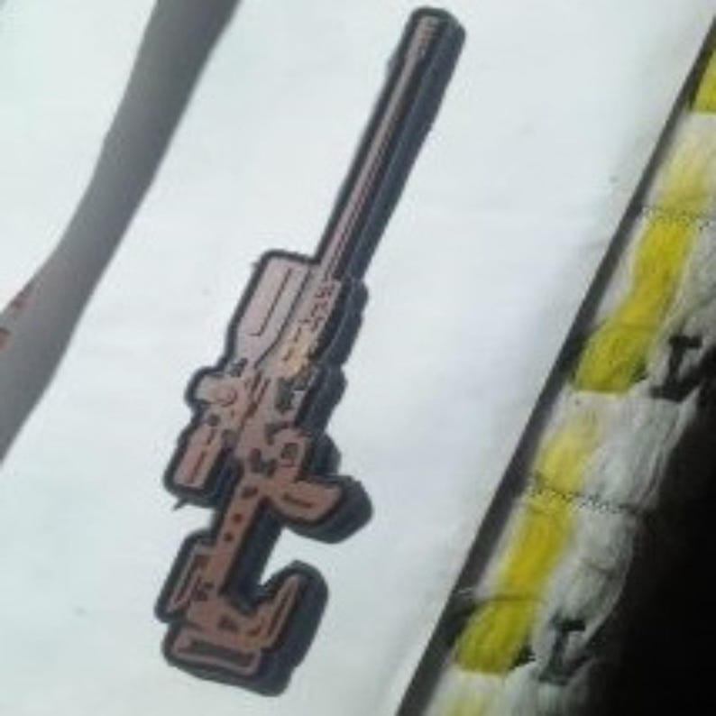 May include: A brown and black gun-shaped patch with a detailed design. The patch is on a white background with a yellow and white striped border.