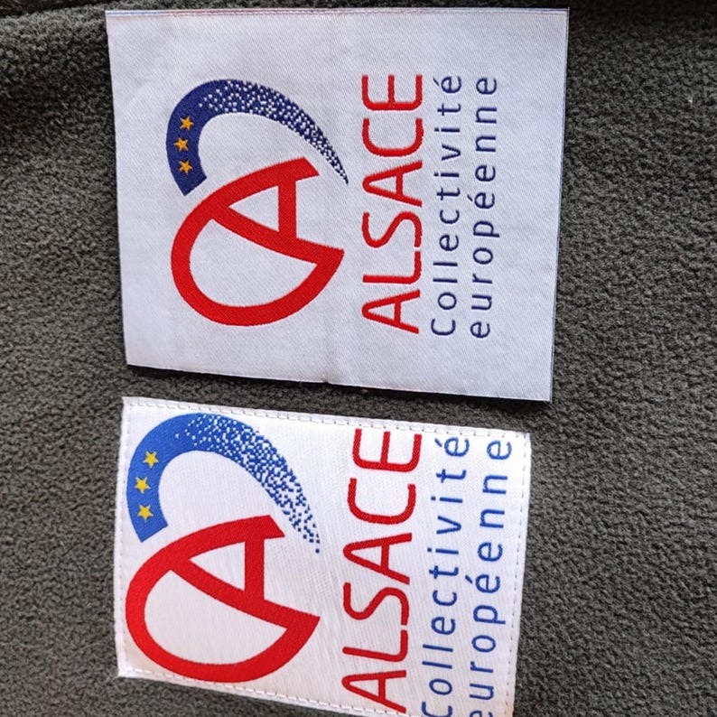 May include: Two white fabric labels with the word "ALSACE" in red, and the text "Collectivit&eacute; europ&eacute;enne" in blue. The labels also feature a red "A" and a blue and yellow design.
