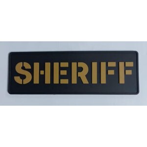 May include: A black rectangular sign with the word "SHERIFF" in gold block letters. The letters have a stencil-like appearance. The sign is likely used for law enforcement identification.