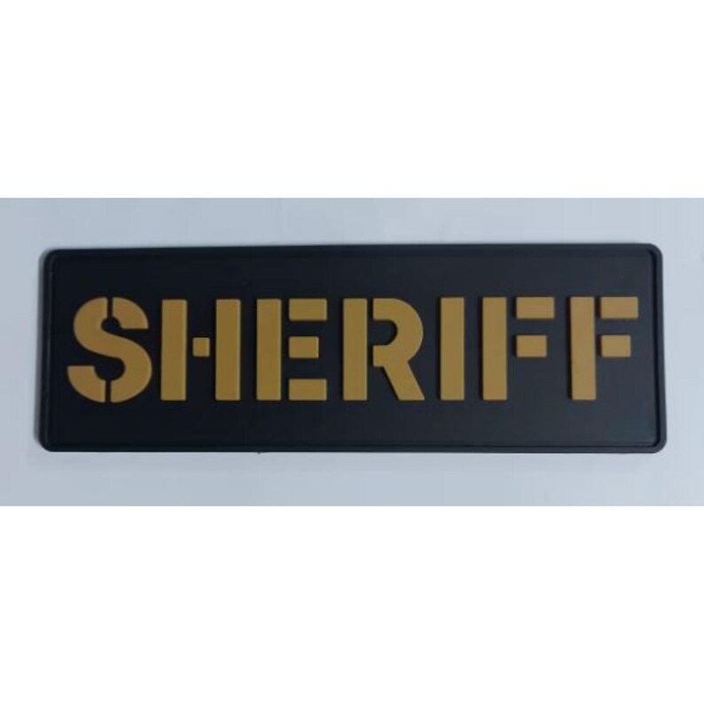 May include: A black rectangular sign with the word "SHERIFF" in gold block letters. The letters have a stencil-like appearance. The sign is likely used for law enforcement identification.