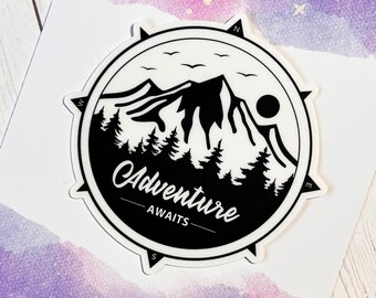 May include: A circular vinyl sticker featuring a black and white mountain landscape with pine trees, a sun, and birds inside a compass frame. The text reads Adventure AWAITS. It is displayed on a purple and white card labeled Vinyl Sticker.