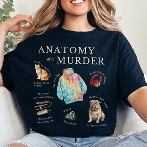 May include: Black t-shirt with a graphic print of a cat, a tie-dye hoodie, a pair of headphones, a dog, and a green gem. The text "Anatomy of a Murder" is printed in white on the shirt.