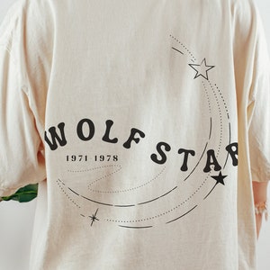 May include: A cream colored t-shirt with a black graphic of a shooting star with the text "Wolfstar 1971-1978" on the back.