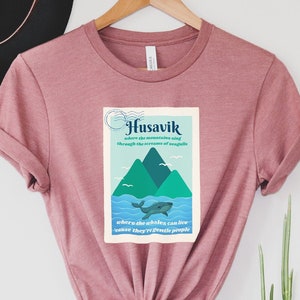 May include: A pink t-shirt with a graphic design featuring a whale and mountains. The graphic text reads "Husavik where the mountains sing through the echoes of whales and they try to drown people".