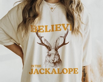 May include: A white oversized t-shirt with a brown and white illustration of a jackalope with antlers. The text "BELIEVE IN THE JACKALOPE" is printed in orange on the shirt.
