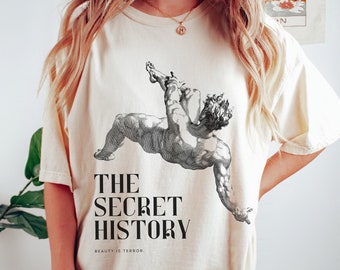 Puede incluir: Una camiseta de color crema con un gráfico en blanco y negro de un cuerpo de hombre. El texto "THE SECRET HISTORY" está impreso debajo del gráfico. El texto "BEAUTY IS TERROR." está impreso debajo del texto.