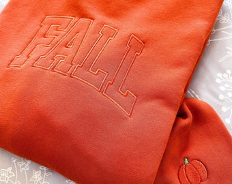 Puede incluir: Una sudadera de color naranja quemado con la palabra "FALL" bordada en una fuente grande de color blanco. Una pequeña calabaza bordada se encuentra en la manga.