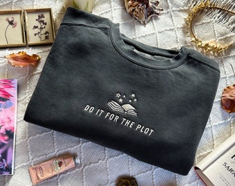 Pode incluir: Camisola cinza escuro com a frase bordada "DO IT FOR THE PLOT" acima de um livro aberto e estrelas. A imagem inclui livros, uma vela, uma concha e objetos decorativos, criando um ambiente acolhedor e literário.