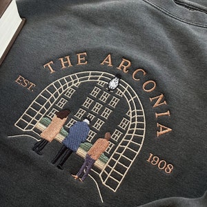 May include: A dark gray sweatshirt with an embroidered design featuring a building with windows, a clock tower, and four people standing in front of it. The text "THE ARCONIA EST. 1908" is embroidered above the building.