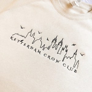 May include: A beige sweatshirt with a black embroidered design of a city skyline and the text "Ketterdam Crow Club".