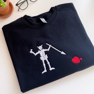 Pode incluir: Camisola de moletom preta com um esqueleto bordado em branco segurando uma lança e um coração vermelho.