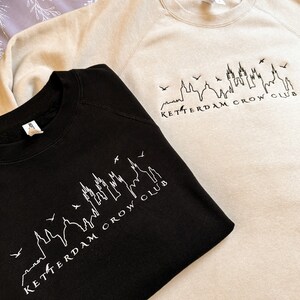 May include: Two sweatshirts, one black and one beige, with a white outline of a city skyline and the text "Ketterdam Crow Club".