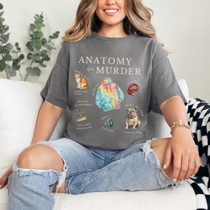 May include: A woman wearing a gray t-shirt with the text "Anatomy of a Murder" and illustrations of a cat, a tie-dye hoodie, headphones, a plane, a ring, and a dog. She is sitting on a white couch with a black and white checkered throw blanket.