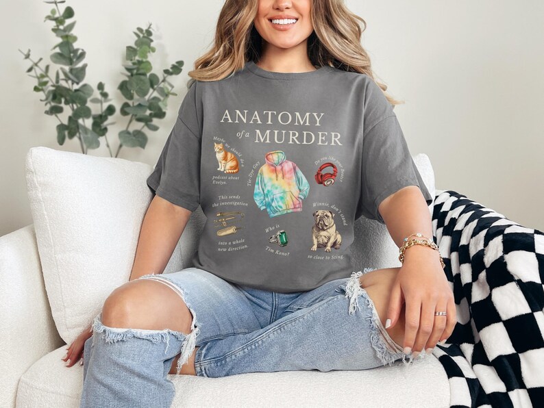 May include: A woman wearing a gray t-shirt with the text "Anatomy of a Murder" and illustrations of a cat, a tie-dye hoodie, headphones, a plane, a ring, and a dog. She is sitting on a white couch with a black and white checkered throw blanket.