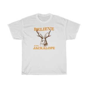 Pu&ograve; includere: T-shirt bianca con un'illustrazione di jackalope marrone e il testo "Believe in the Jackalope" in arancione.