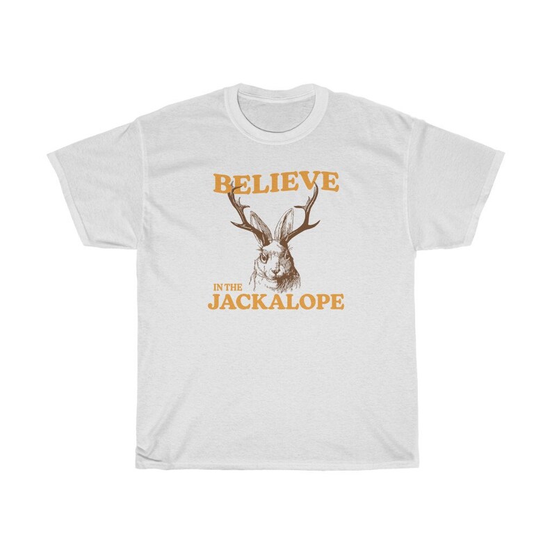 Pu&ograve; includere: T-shirt bianca con un'illustrazione di jackalope marrone e il testo "Believe in the Jackalope" in arancione.