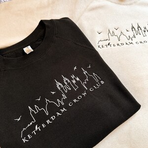 May include: Two sweatshirts with a skyline graphic and the text "Rotterdam Crow Club". The sweatshirt on the top is beige and the sweatshirt on the bottom is black.