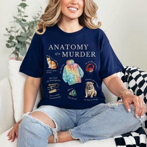 May include: A navy blue t-shirt with the text "Anatomy of a Murder" printed on the front. The shirt features illustrations of a cat, a tie-dye hoodie, a crab, a dog, a pair of hair clippers, and a green gem.