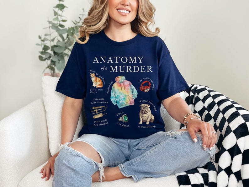 May include: A navy blue t-shirt with the text "Anatomy of a Murder" printed on the front. The shirt features illustrations of a cat, a tie-dye hoodie, a crab, a dog, a pair of hair clippers, and a green gem.