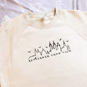 May include: A beige sweatshirt with a black outline of a city skyline and the text "Ketterdam Crow Club".