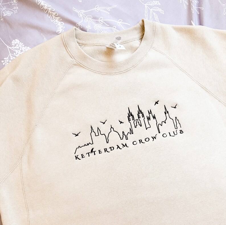 May include: A beige sweatshirt with a black outline of a city skyline and the text "Ketterdam Crow Club".