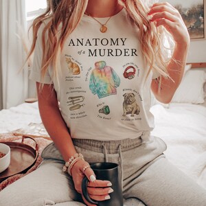 May include: White t-shirt with the text "Anatomy of a Murder" and illustrations of a cat, a tie-dye hoodie, headphones, a bulldog, and a wrench. The text on the shirt includes the phrases "Tie-Dye Gay", "podcast about Evelyn", "This sends the investigation into a whole new direction", "Who is Tim Kono?", and "Winnie, don't stand so close to Sting."