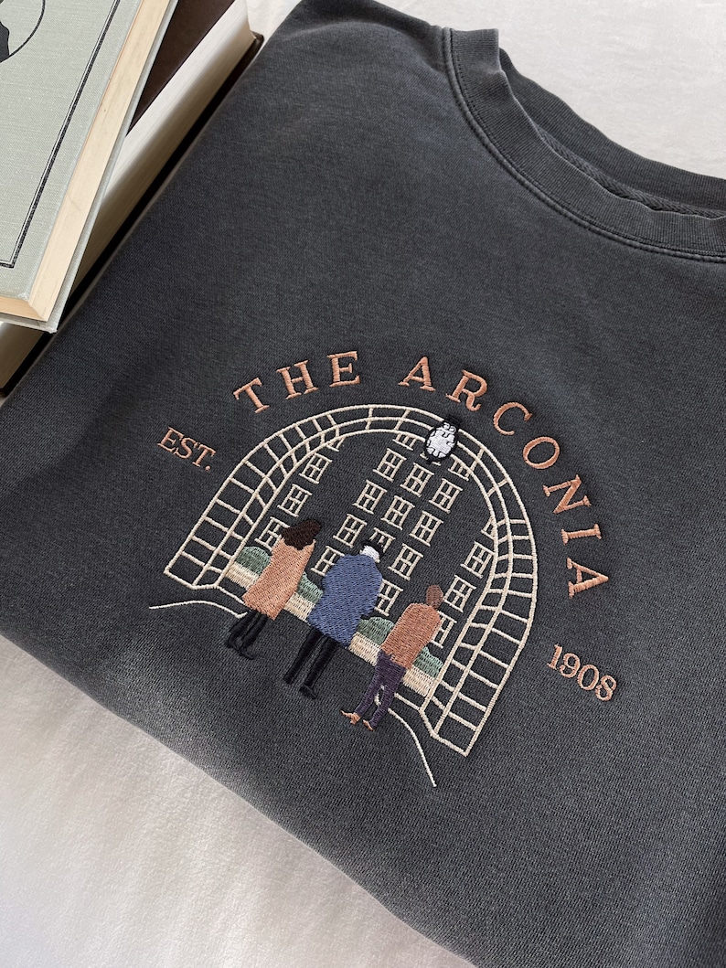 May include: A dark gray sweatshirt with an embroidered design featuring a building with an archway, the text "THE ARCONIA EST. 1908", and four people standing in front of the building.
