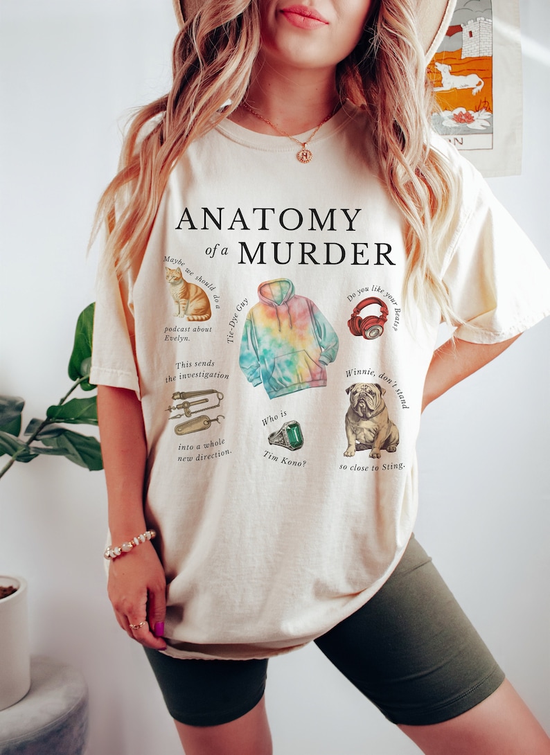 May include: A light beige t-shirt with the text "Anatomy of a Murder" printed in black. The shirt features several illustrations, including a tie-dye hoodie, a cat, a pair of headphones, a bulldog, and a ring.