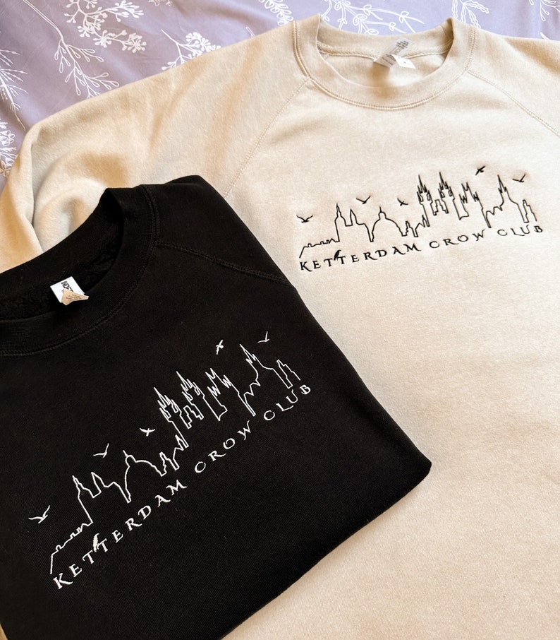 May include: Two sweatshirts, one black and one beige, with a white outline of a city skyline and the text "Ketterdam Crow Club".
