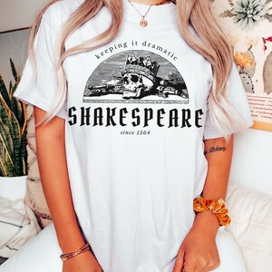 May include: White t-shirt featuring a black and white graphic of a skull wearing a crown. The graphic is in a circle with the text "keeping it dramatic" above the skull and "SHAKESPEARE since 1564" below the skull.