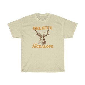 Pu&ograve; includere: Una maglietta beige con il testo "BELIEVE IN THE JACKALOPE" in arancione. Sopra il testo c'&egrave; un disegno di un jackalope con le corna.
