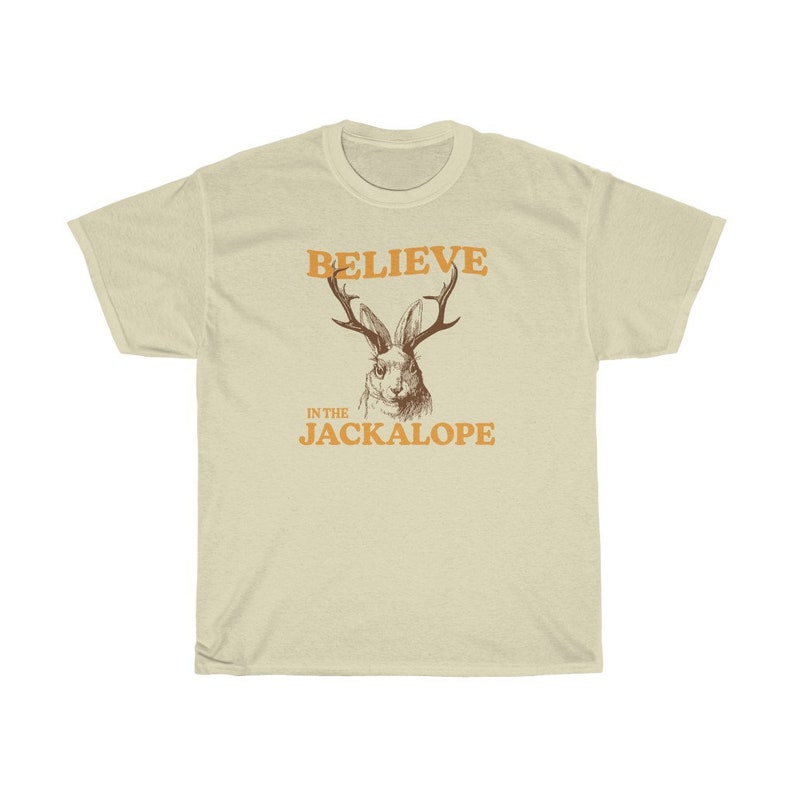 Pu&ograve; includere: Una maglietta beige con il testo "BELIEVE IN THE JACKALOPE" in arancione. Sopra il testo c'&egrave; un disegno di un jackalope con le corna.