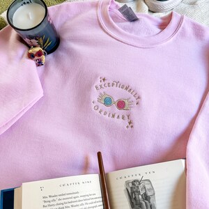 May include: Pink sweatshirt with a gold embroidered design of a pair of glasses with a lightning bolt and the text "Exceptionally Ordinary". The sweatshirt is laying on a white and green blanket with a candle and a book.
