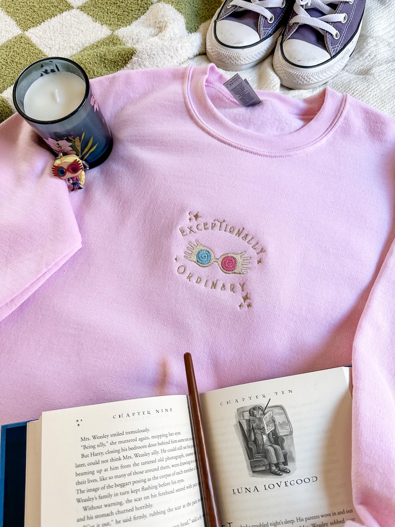 May include: Pink sweatshirt with a gold embroidered design of a pair of glasses with a lightning bolt and the text "Exceptionally Ordinary". The sweatshirt is laying on a white and green blanket with a candle and a book.