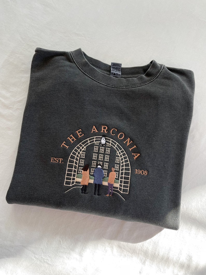 May include: A dark gray sweatshirt with an embroidered design of a building with an archway and three people standing in front of it. The text "THE ARCONIA EST. 1908" is embroidered around the design.