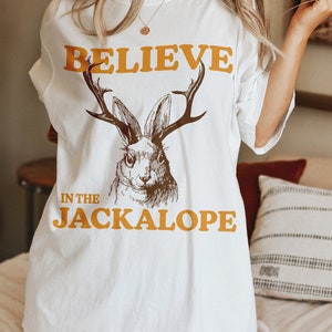 Pu&ograve; includere: Maglietta bianca con un'illustrazione marrone e bianca di un jackalope, un coniglio con le corna. Il testo "BELIEVE IN THE JACKALOPE" &egrave; stampato in arancione sulla maglietta.